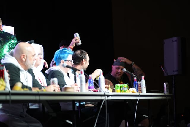 A group of people sitting at a table on a stage, engaged in a discussion or panel, with various drinks and items in front of them