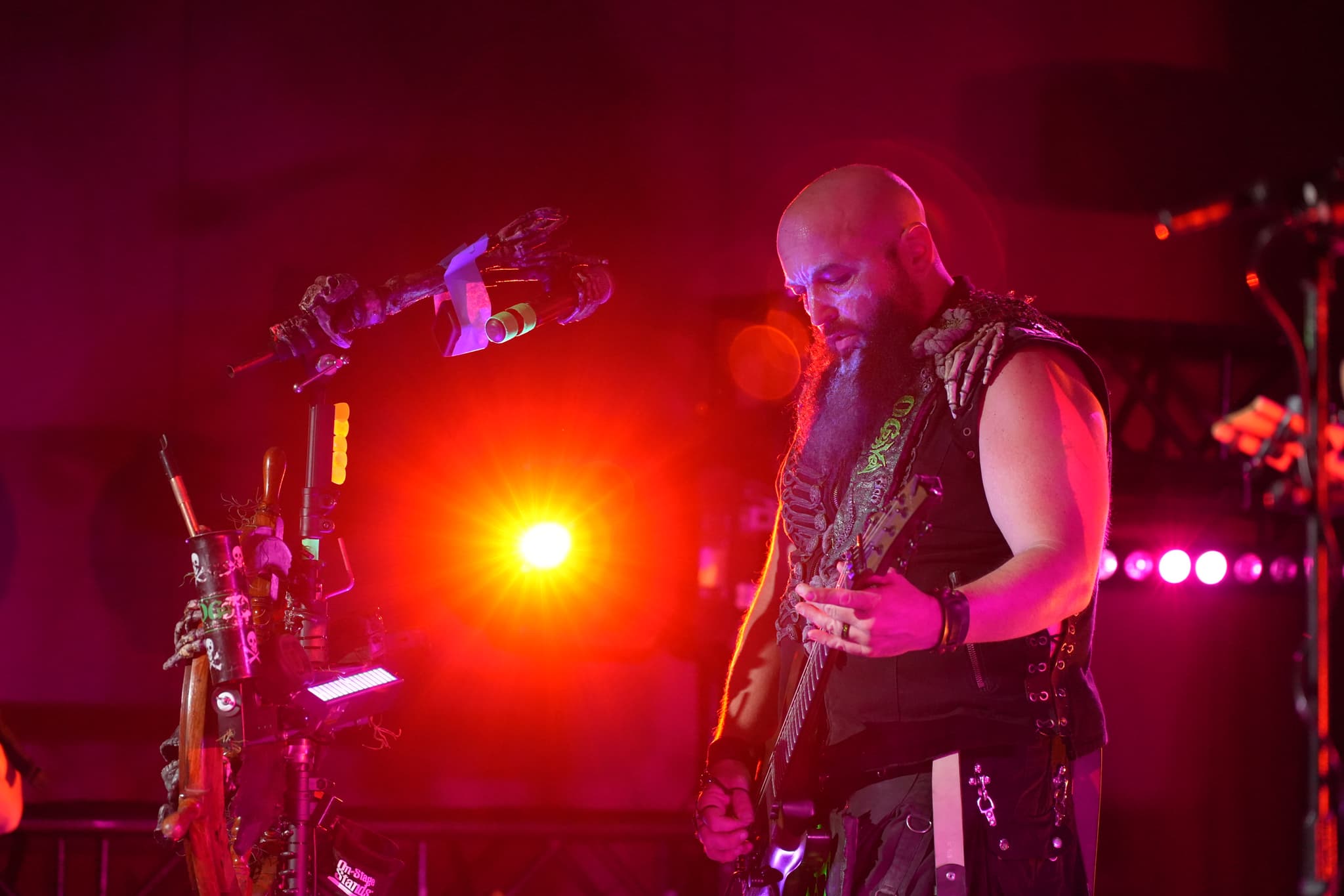 A musician with a bald head and a beard plays an electric guitar on stage, illuminated by vibrant red and purple lights