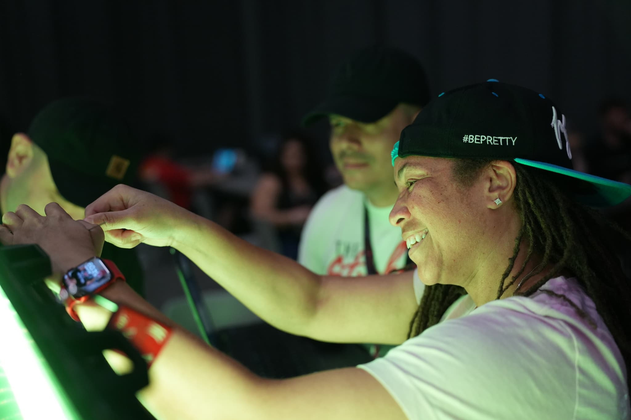 Three people interacting with a glowing object, with one person smiling and pointing at it. They are in a dimly lit environment, and all are wearing casual clothing and caps