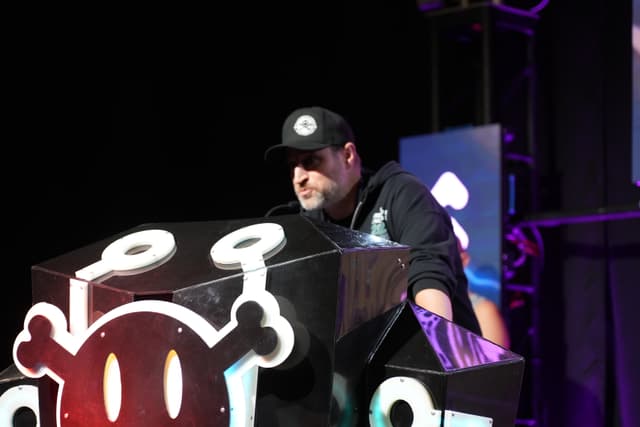 A person wearing a black cap and hoodie speaking at a podium decorated with a stylized skull graphic, with a dark background and colorful lighting