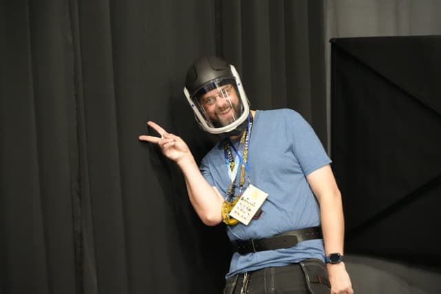 A person wearing a helmet and a blue shirt, making a peace sign gesture, with a lanyard and badge around their neck, standing in front of a dark curtain