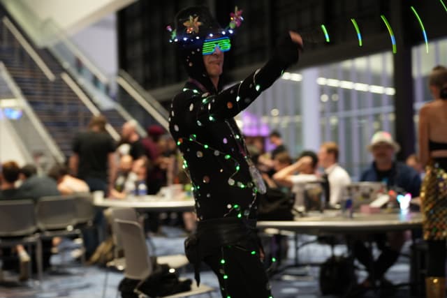 A person wearing a black suit with colorful lights and a headpiece, standing in a large room with people seated at tables in the background