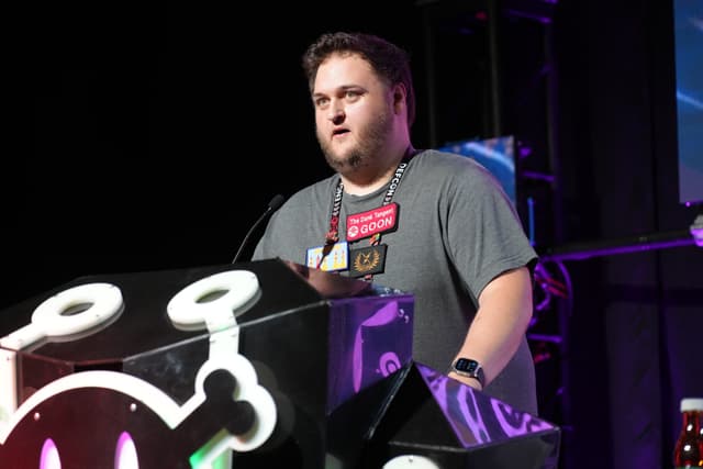 A man stands at a podium with a microphone, wearing a gray shirt and a badge around his neck, speaking at an event. The podium has a design featuring a stylized skull and crossbones