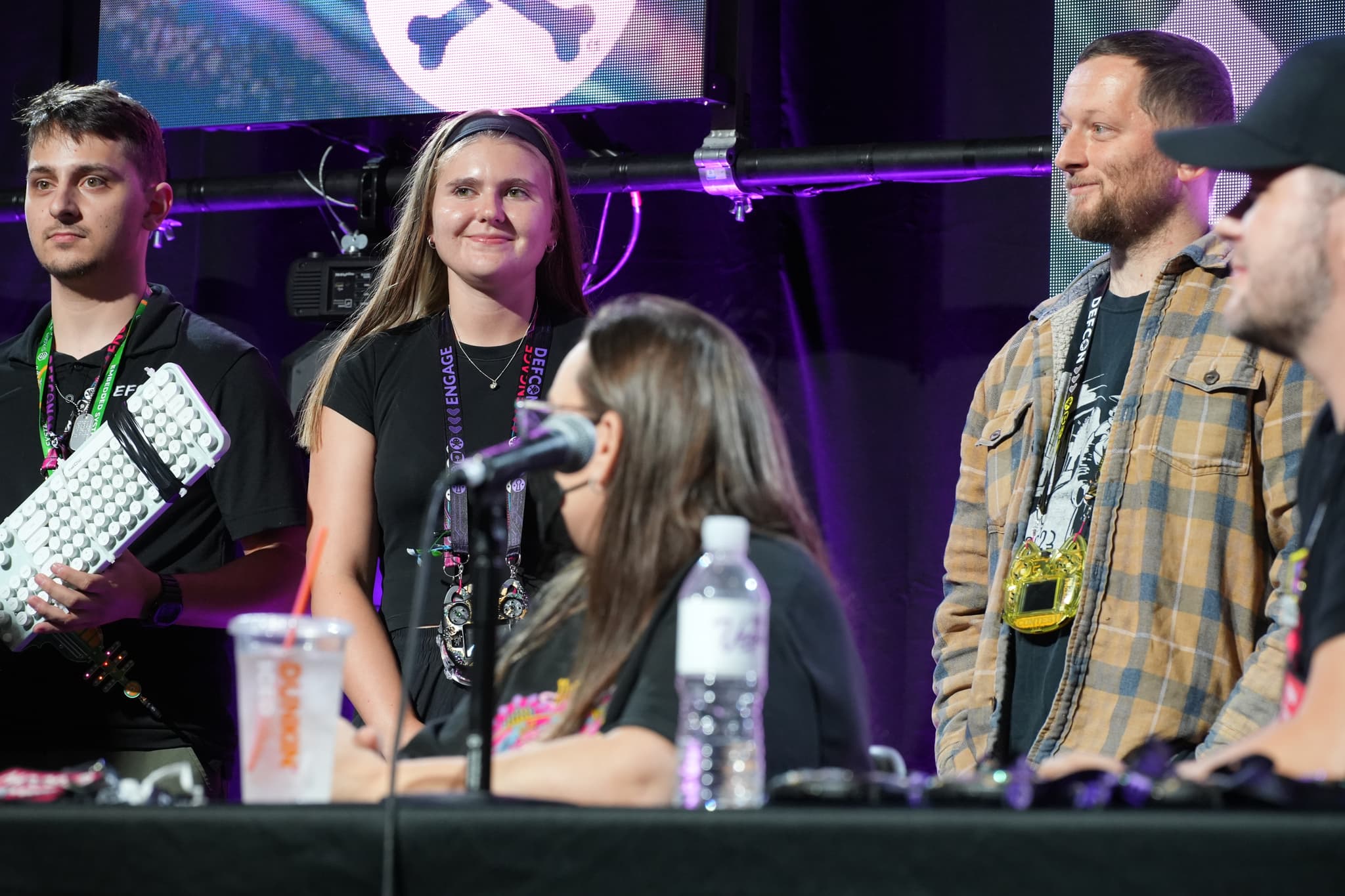 A group of people standing and sitting at a panel, with microphones and drinks on the table, under purple lighting