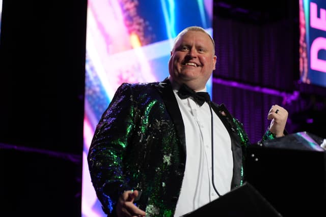 A person wearing a sparkly green and black jacket with a white shirt and black bow tie, standing on a stage with colorful lights in the background