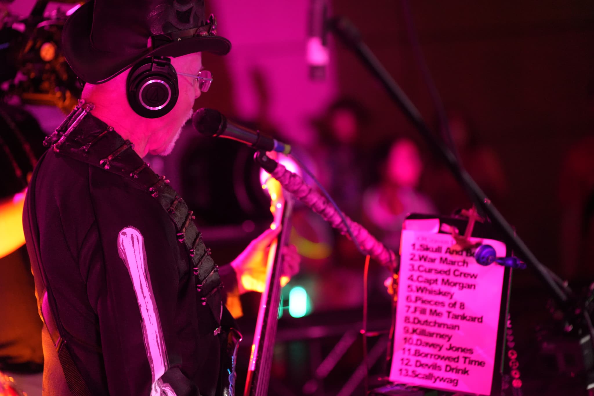 A musician wearing headphones and a hat performs on stage, with a microphone and a setlist visible in the foreground. The background is illuminated with pink and purple lighting, and an audience is faintly visible