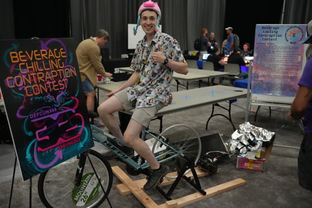 A person wearing a pink helmet and floral shirt is sitting on a stationary bicycle, giving a thumbs-up. A colorful sign nearby reads Beverage Cooling Contraption Contest. Other people are in the background, some working at tables