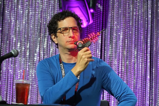 A person wearing glasses and a blue shirt is holding a microphone while sitting at a table with a drink, in front of a sparkly curtain backdrop