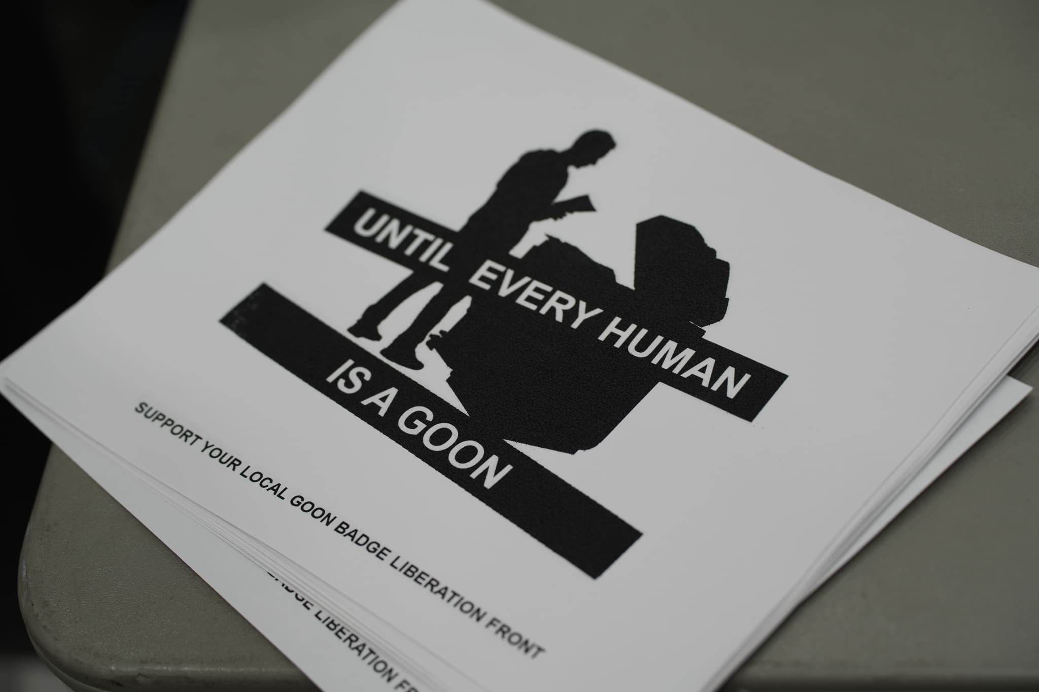 A stack of papers with a black and white graphic depicting a person and a large rock, accompanied by the text UNTIL EVERY HUMAN IS A GOON. Additional text at the bottom reads SUPPORT YOUR LOCAL GOON SQUAD LABORATORY FRONT