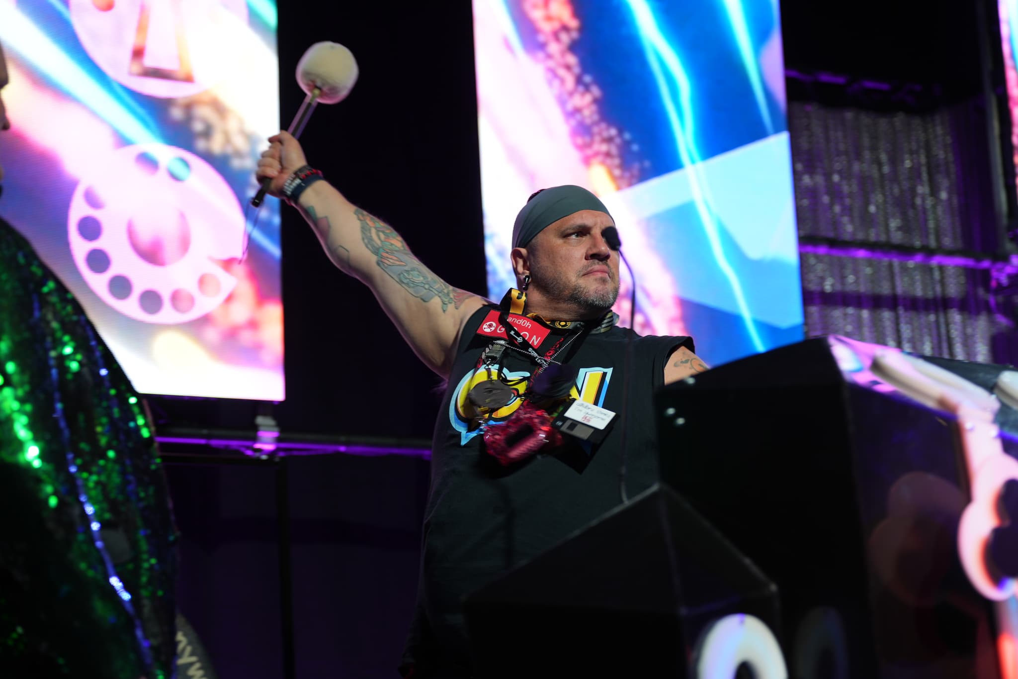 A person wearing a sleeveless shirt and a beanie, holding a mallet, stands on a stage with colorful, abstract backgrounds