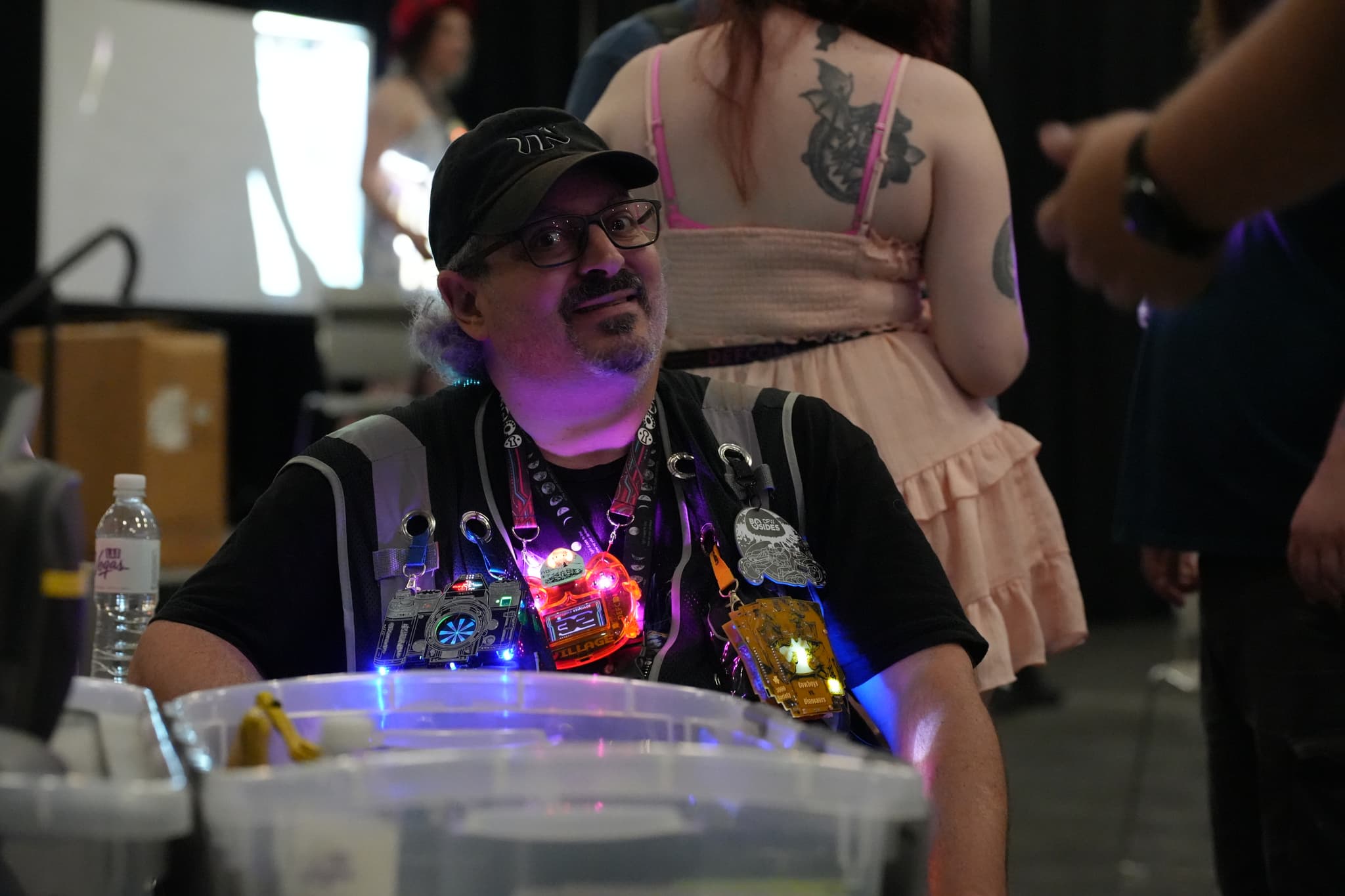 A man wearing a cap and glasses, adorned with colorful, glowing accessories, sits behind a table with various items In the background, a person with a tattoo on their back is visible, along with other people and a whiteboard