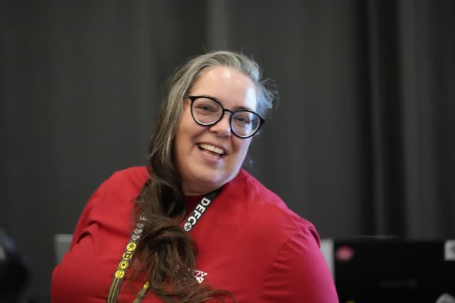 A person with long hair and glasses, wearing a red shirt and a lanyard, smiling in front of a dark background