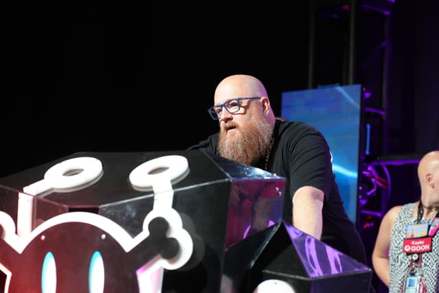 A bearded man with glasses speaking at a podium with a stylized design, while another person stands in the background