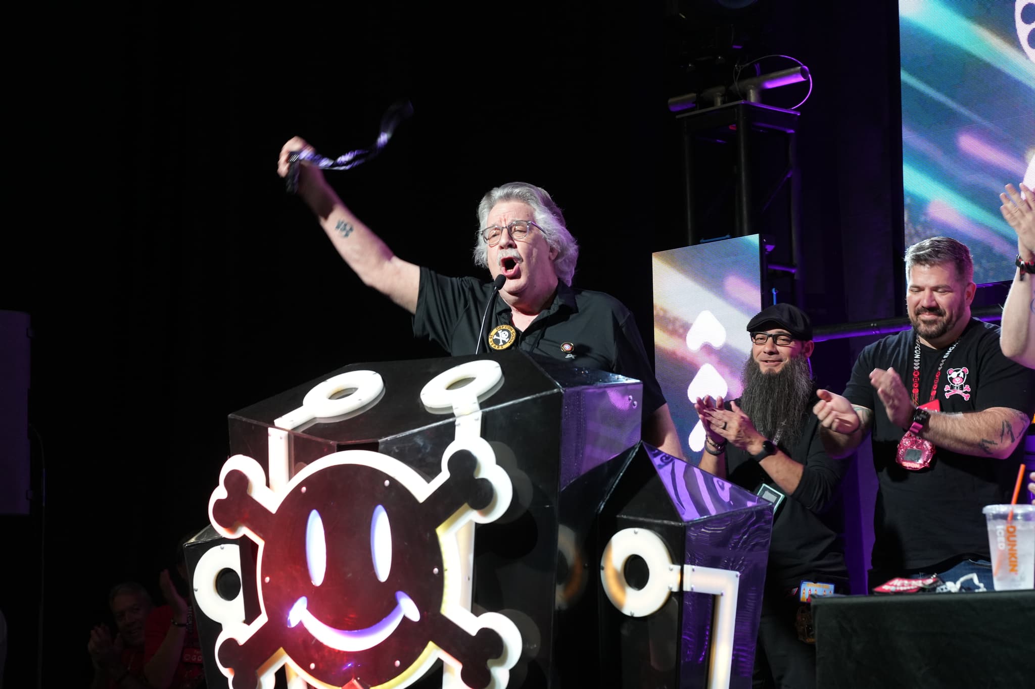 A person standing behind a podium with a smiley face design, raising an object in the air, while others nearby applaud