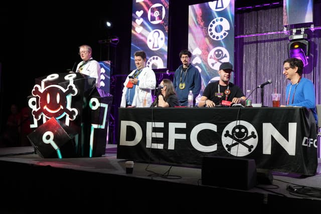 A group of people on a stage at DEFCON, with one person speaking at a podium and others seated at a table with the DEFCON logo. The background features illuminated screens and technical equipment