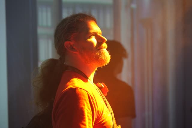 A person with a beard and long hair tied back, wearing an orange shirt, stands in profile with eyes closed, bathed in warm sunlight