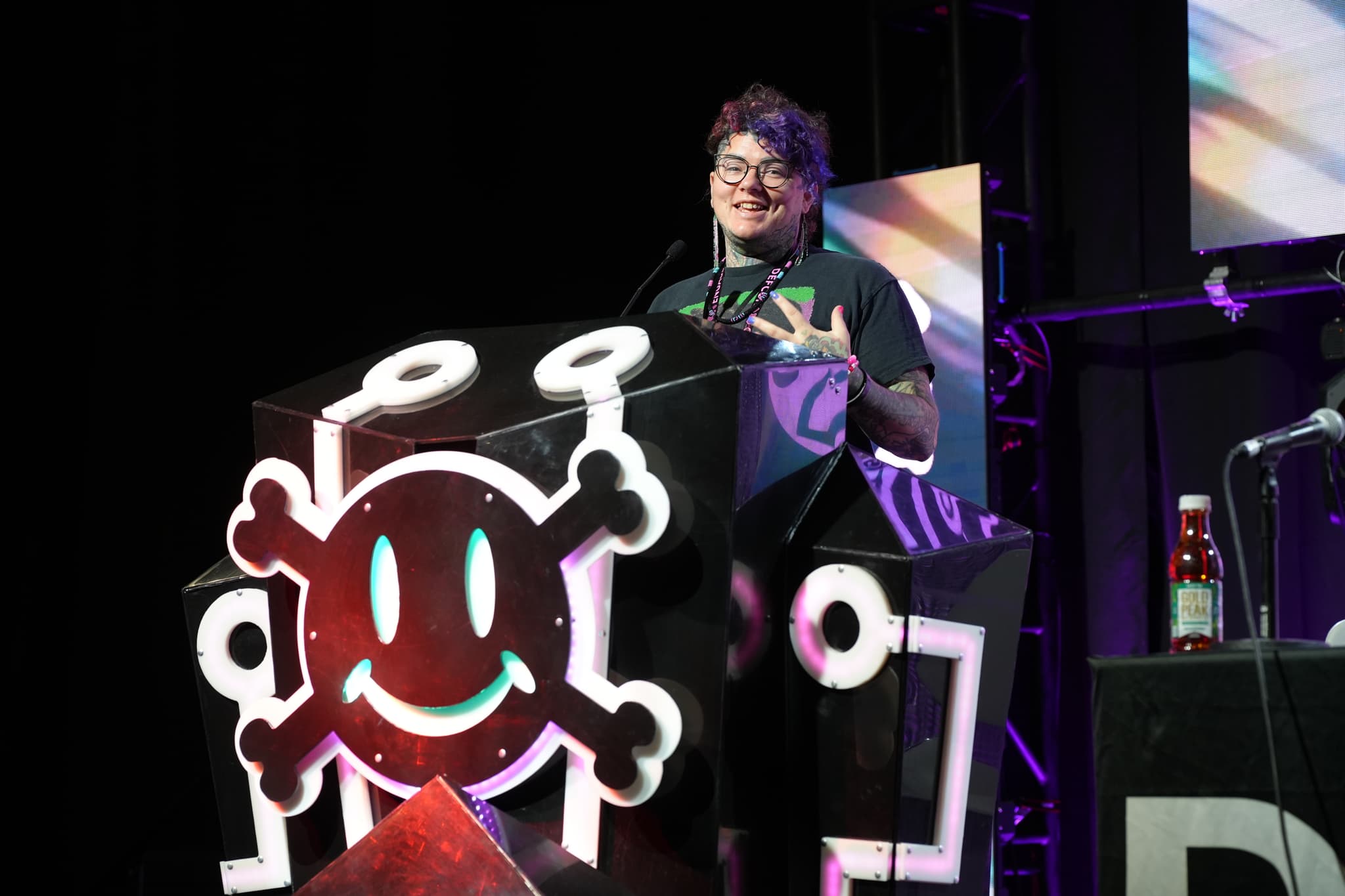 A person standing behind a podium decorated with a large, stylized skull and crossbones symbol, speaking into a microphone on a stage