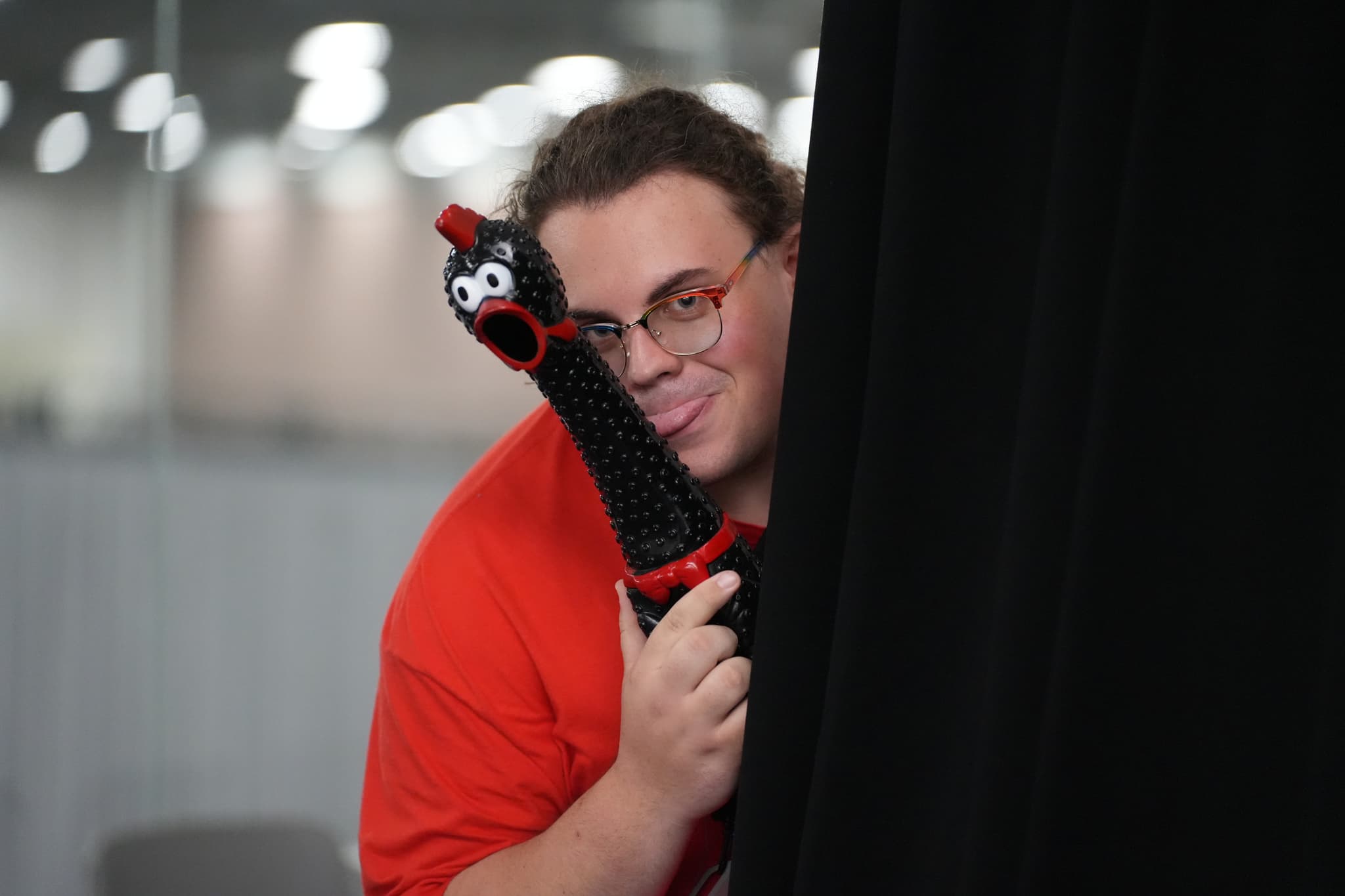 A person in a red shirt peeks from behind a black curtain, holding a black object with googly eyes and a red mouth