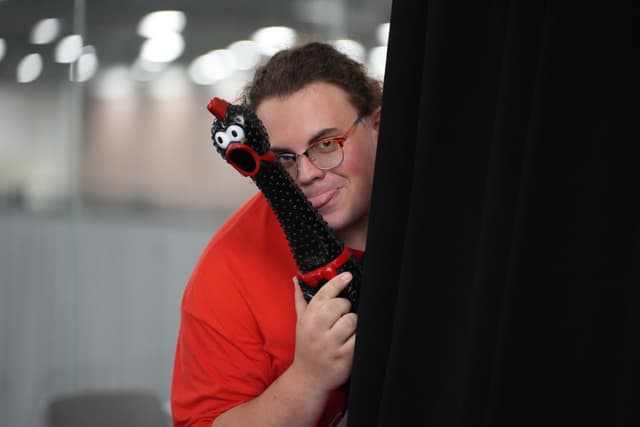 A person in a red shirt peeks from behind a black curtain, holding a black object with googly eyes and a red mouth