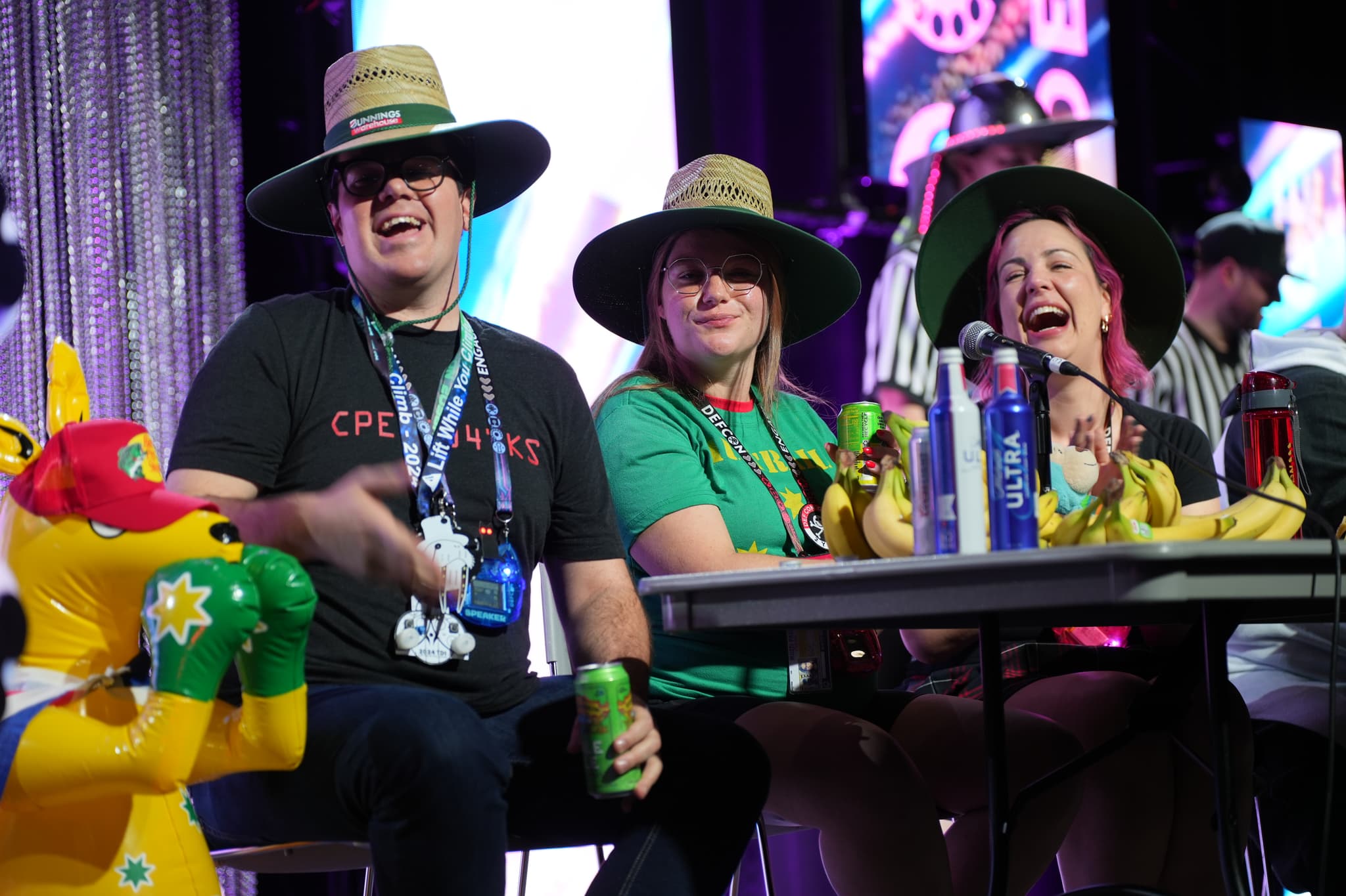 Three people wearing wide-brimmed hats sitting at a table with drinks and inflatable kangaroos, smiling and enjoying themselves in a festive environment