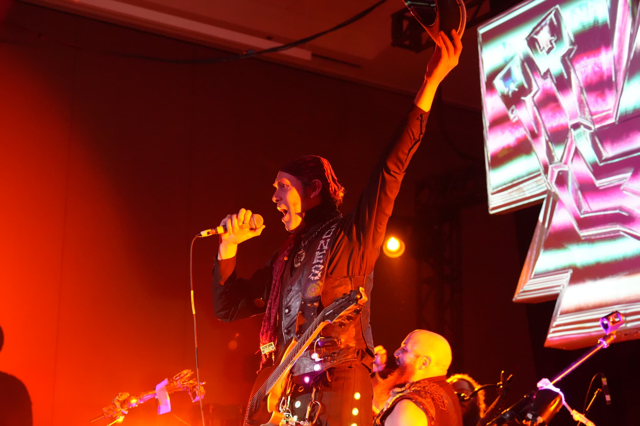 A musician on stage passionately singing into a microphone with one arm raised, accompanied by a band member playing an instrument, and vibrant, colorful lighting and visuals in the background