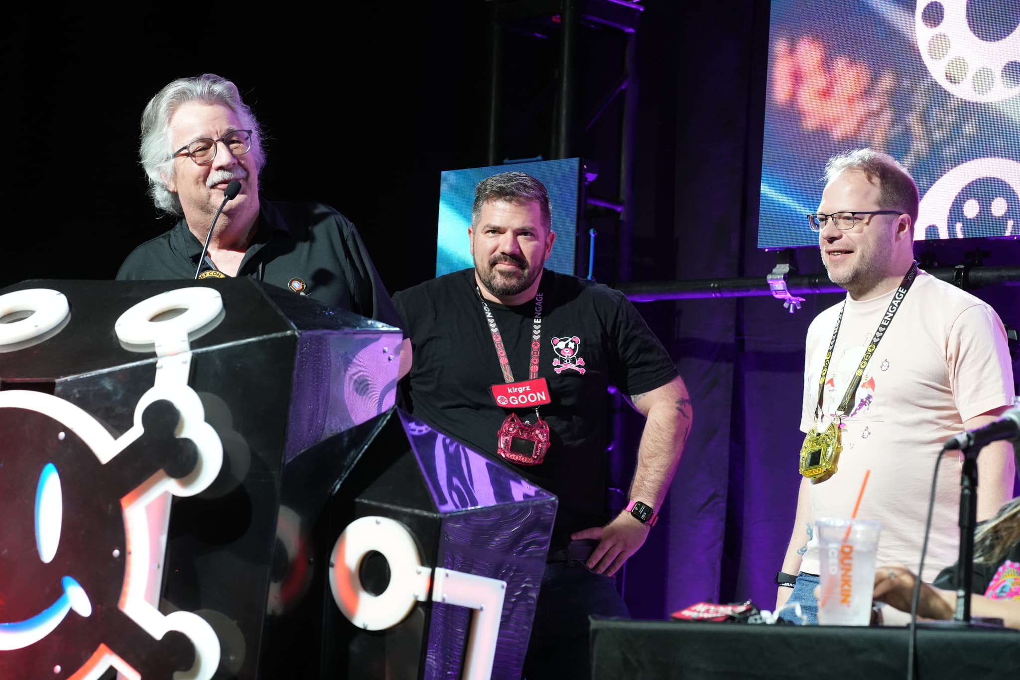 Three men standing on a stage, with one speaking into a microphone. The stage is decorated with a large black and white design featuring a skull and crossbones motif. The background includes colorful lighting and a screen displaying abstract graphics