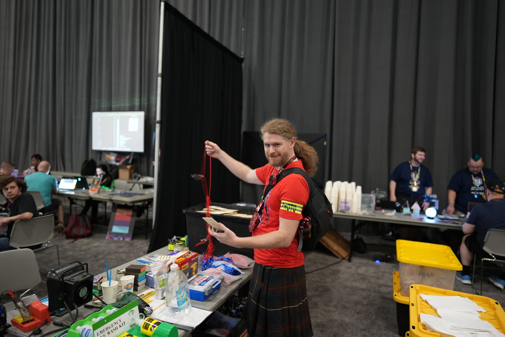 A person with long hair, wearing a red shirt and a kilt, is holding a long red object in a room filled with various items and people working at tables