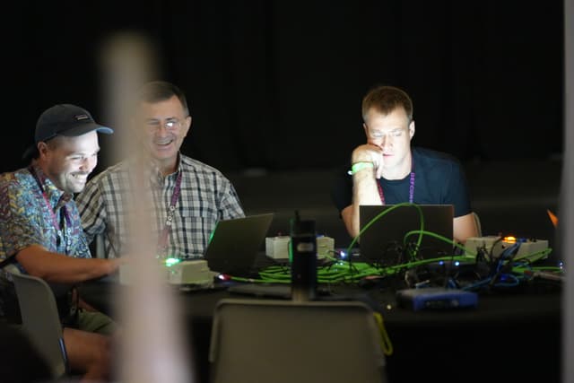 Three people sitting at a table with laptops, engaged in a discussion or collaborative work, with a dark background and some electronic equipment visible on the table