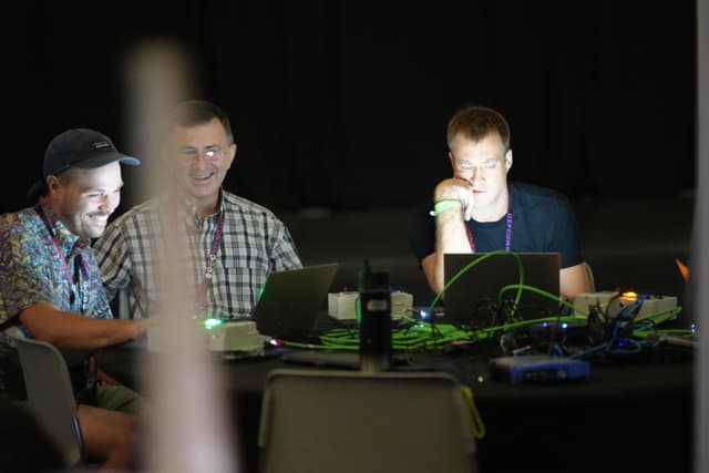 Three people sitting at a table with laptops, engaged in a discussion or collaborative work, with a dark background and some electronic equipment visible on the table