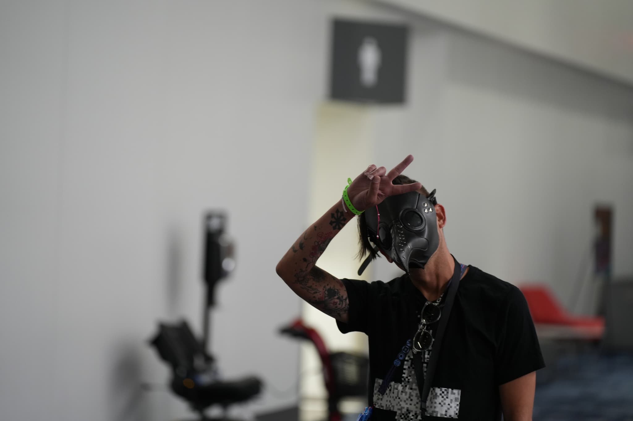 A person wearing a black T-shirt and a mask is posing with one hand making a gesture near their head. The background includes a blurred indoor setting with a sign indicating a restroom