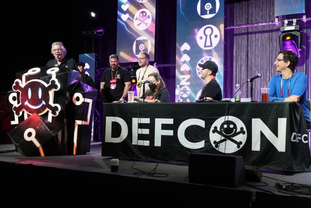 A panel of five individuals on a stage at DEFCON, with one person speaking at a podium and the others seated behind a table with a DEFCON banner. The background features various tech-themed decorations and lighting