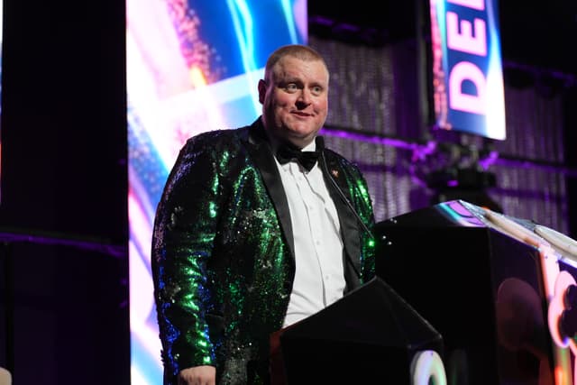 A man in a shiny green jacket and bow tie stands at a podium with colorful lights and signs in the background