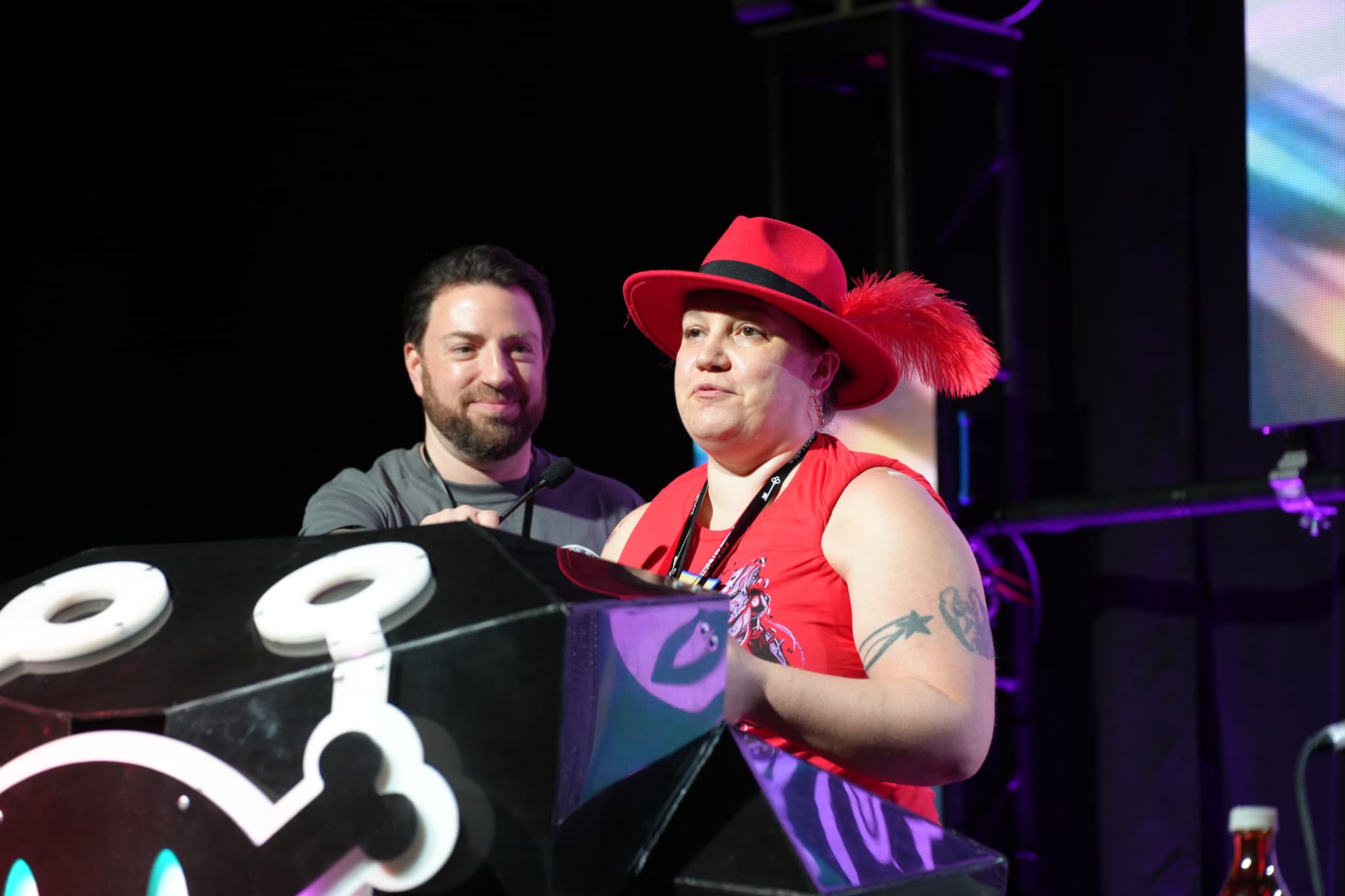 Two individuals standing behind a podium, with one wearing a red hat and red outfit, and the other in a dark shirt. The podium has a design featuring a white skull and crossbones