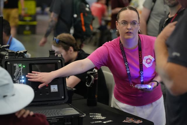 A person in a pink shirt is demonstrating equipment to a group of people at an indoor event