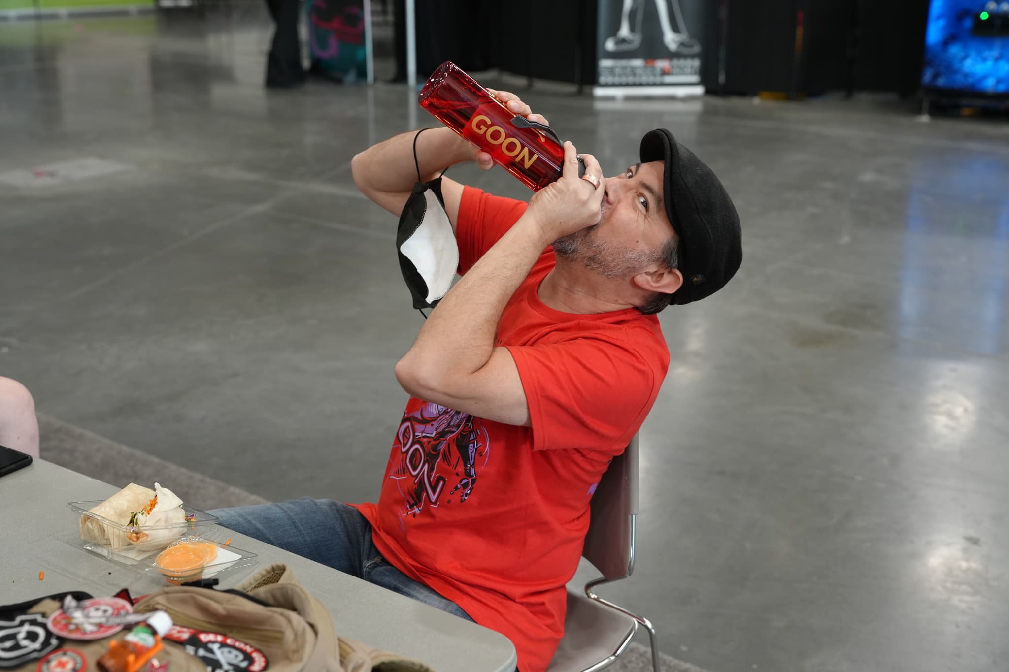 A person in a red shirt and black cap is sitting at a table, drinking from a large red bottle. The table has various items on it, including food and a drink. The background appears to be an indoor setting with a concrete floor and some booths or displays