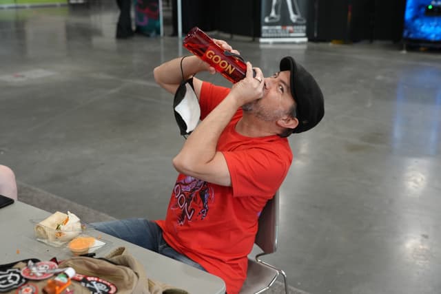 A person in a red shirt and black cap is sitting at a table, drinking from a large red bottle. The table has various items on it, including food and a drink. The background appears to be an indoor setting with a concrete floor and some booths or displays