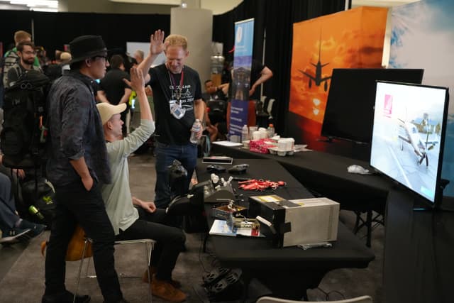 A group of people interacting at a technology booth, with one person sitting and raising their hand, another standing and observing, and a third person holding a drink. A large monitor displays content related to drones, and various equipment and promotional materials are spread across the table