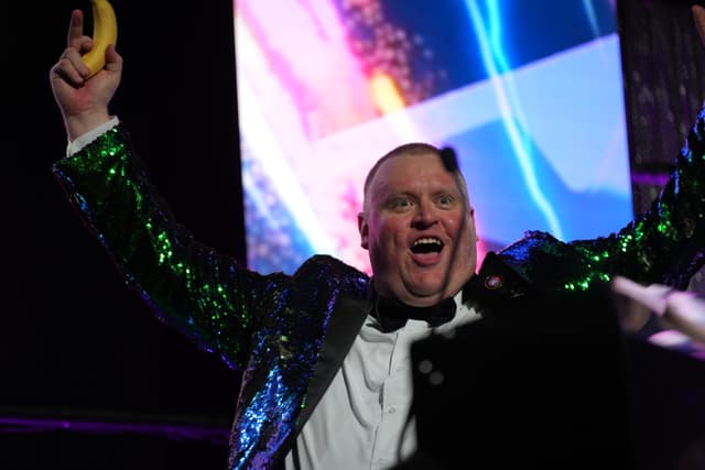 A person wearing a sparkly green jacket and a white shirt, holding a banana in one hand, with a colorful, vibrant background