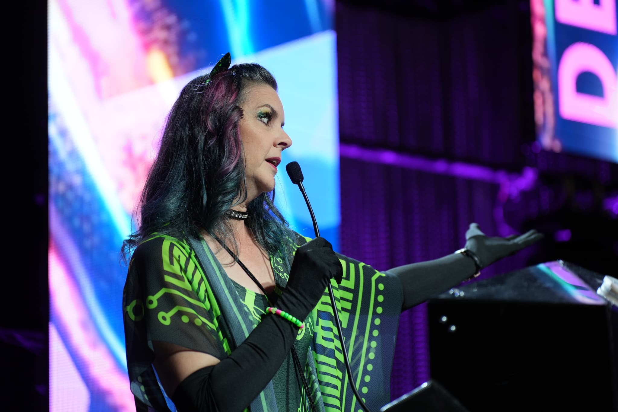 A person with colorful hair and wearing a vibrant outfit speaks into a microphone on stage, gesturing with one hand. The background features bright, abstract lighting and a partial view of a large screen