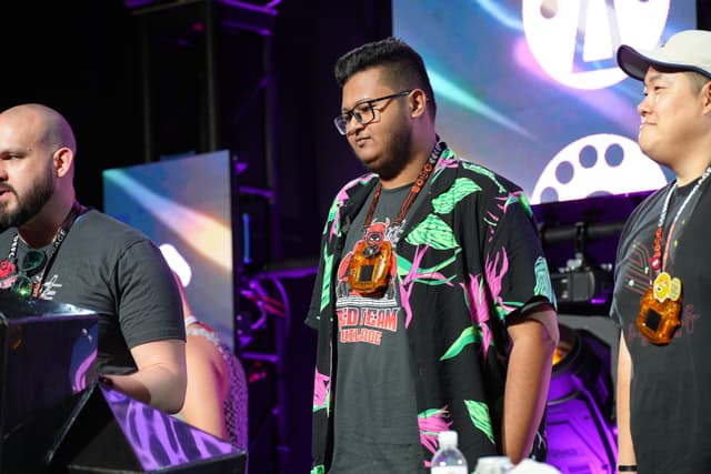 Three men standing on a stage, with one speaking into a microphone. They are wearing casual clothing and large, colorful medallions around their necks. The background features vibrant lighting and a large screen