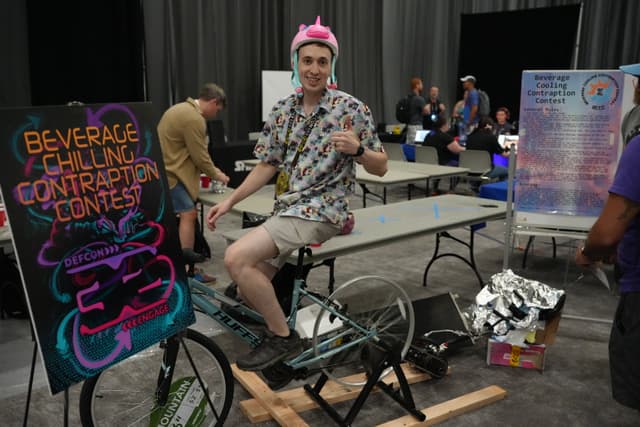 A person wearing a pink helmet and a floral shirt is sitting on a stationary bike, giving a thumbs-up. A colorful sign next to the bike reads Beverage Chillin Contraption Contest. Several people are in the background, and there are tables and various items around the area