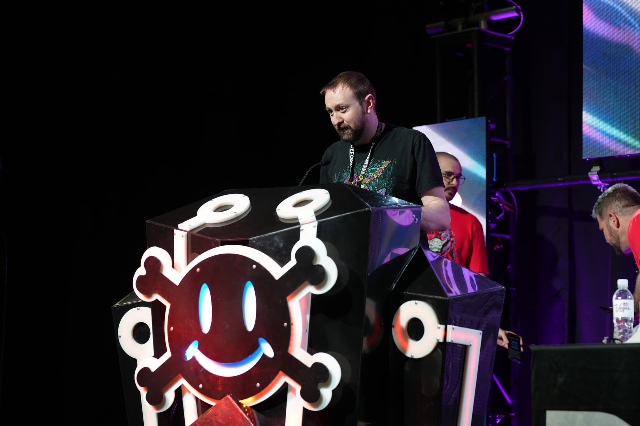 A man standing at a podium decorated with a smiling skull and crossbones, with several people in the background on a stage