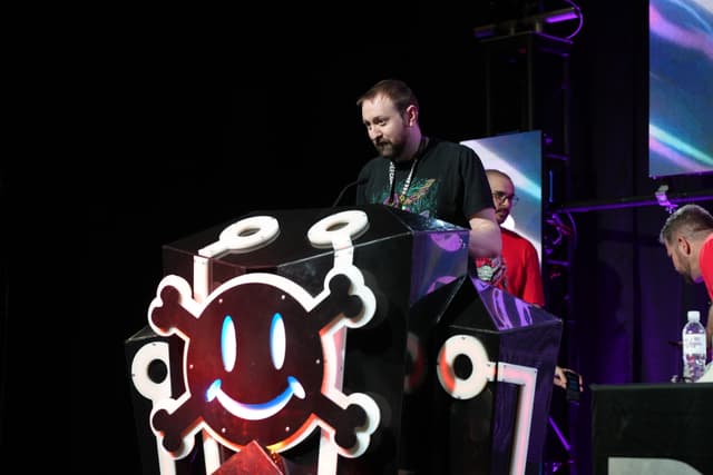 A man standing at a podium decorated with a smiling skull and crossbones, with several people in the background on a stage