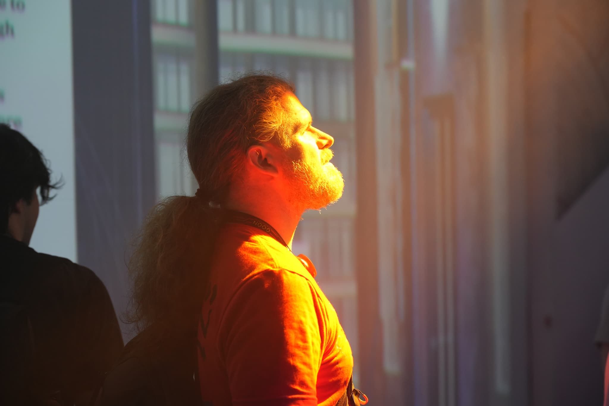 A person with a beard and ponytail, wearing an orange shirt, is bathed in warm light while looking upwards