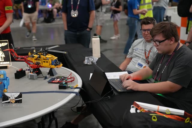 Two individuals working on laptops at a table with various robotics equipment, surrounded by other people in a busy indoor setting