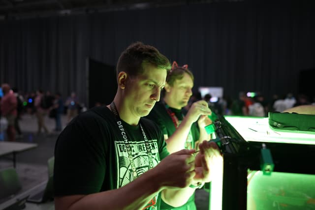 Two individuals are focused on an activity involving a green-lit display in a dimly lit room with other people in the background