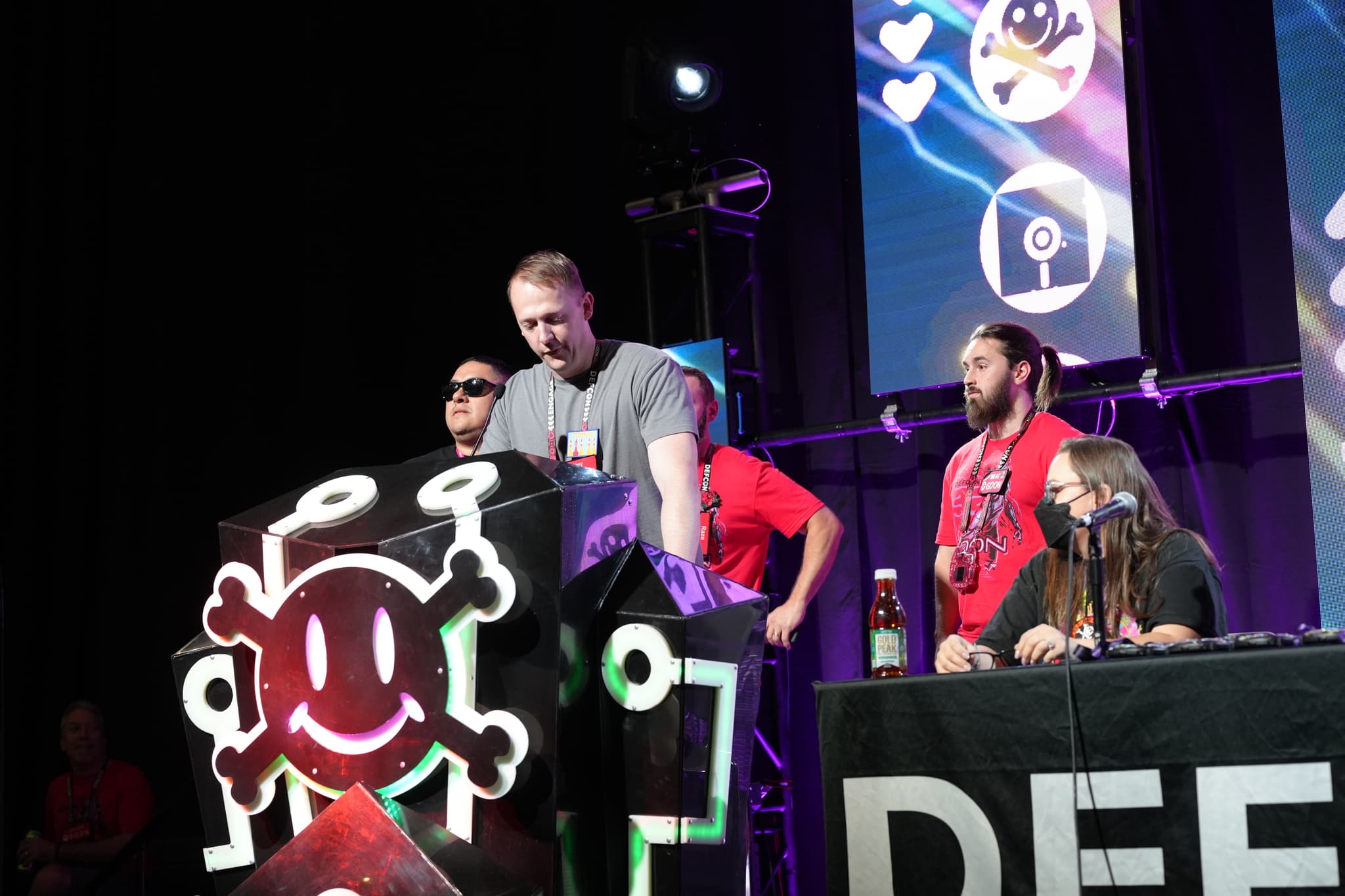 A group of people on a stage, with one person standing behind a podium featuring a skull and crossbones design. Others are seated at a table with the word DEF visible. The background includes screens displaying various symbols