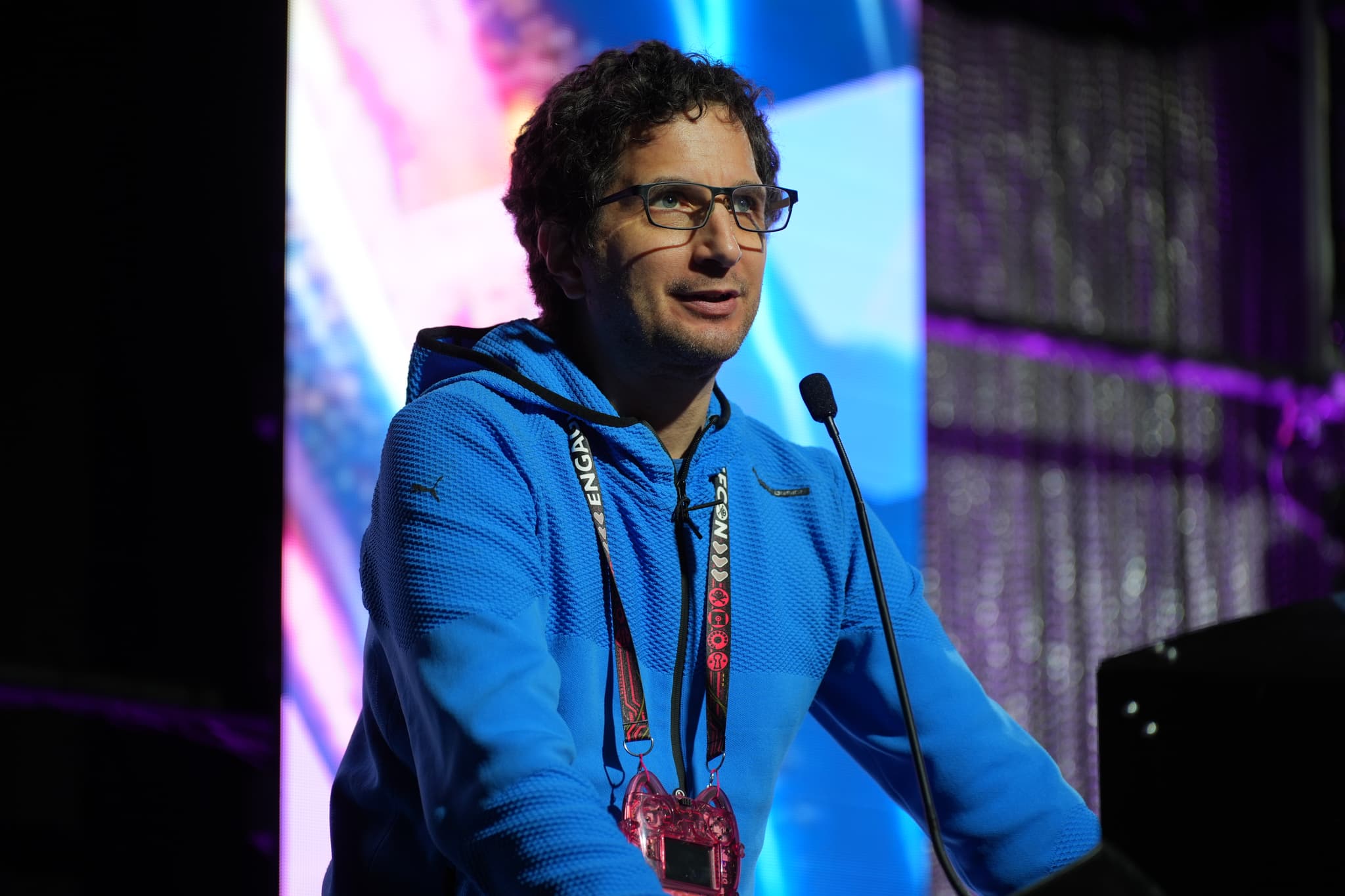 A person wearing a blue hoodie and glasses is speaking into a microphone on a stage with colorful lighting in the background