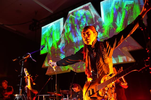 A musician performing on stage with vibrant, colorful projections in the background. The musician is holding a guitar and has their arms outstretched, while other band members play instruments in the background