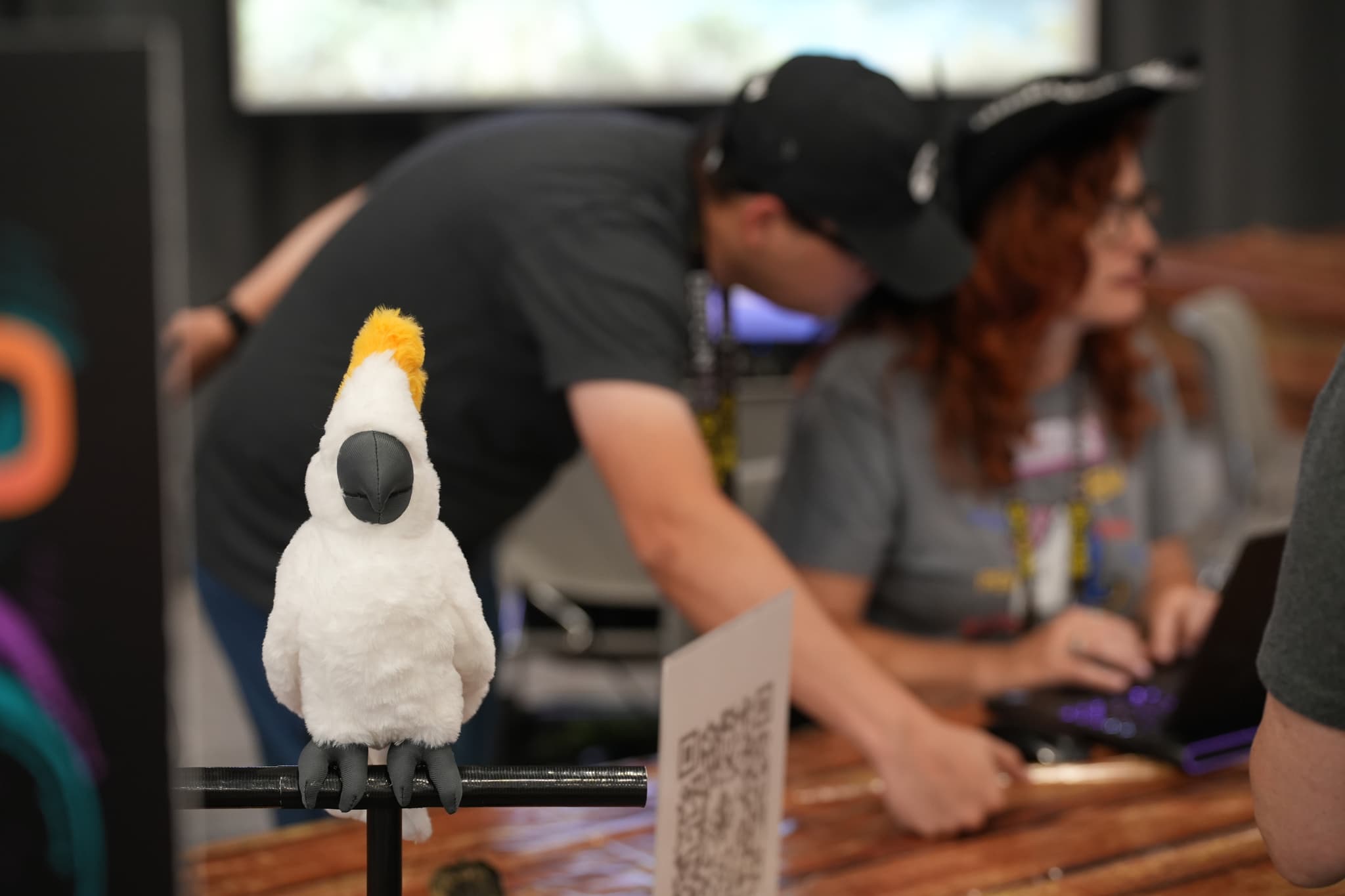 A white cockatoo with a yellow crest perched on a stand, with two people in the background working at a table